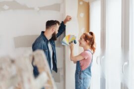 Man standing next to a woman wearing jean overalls holding different color samples in a room undergoing renovations.