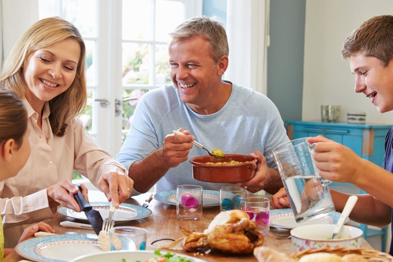 A family sits and smiles at a dinner table. The mom serves the daughter, the dad holds a dish, and the son pours water.
