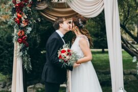 A bride and groom kiss under a floral archway, surrounded by flower petals during their outdoor wedding ceremony.