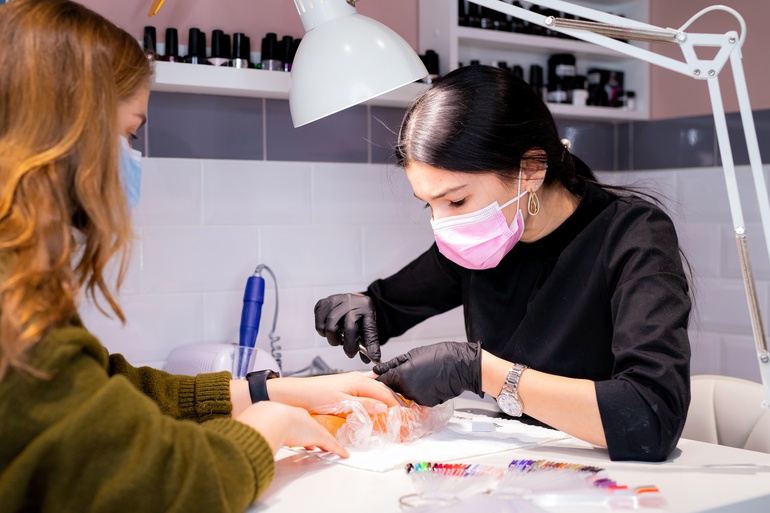 A woman working on another woman's nails. She's wearing a pink mask and filing the customer's nails down.