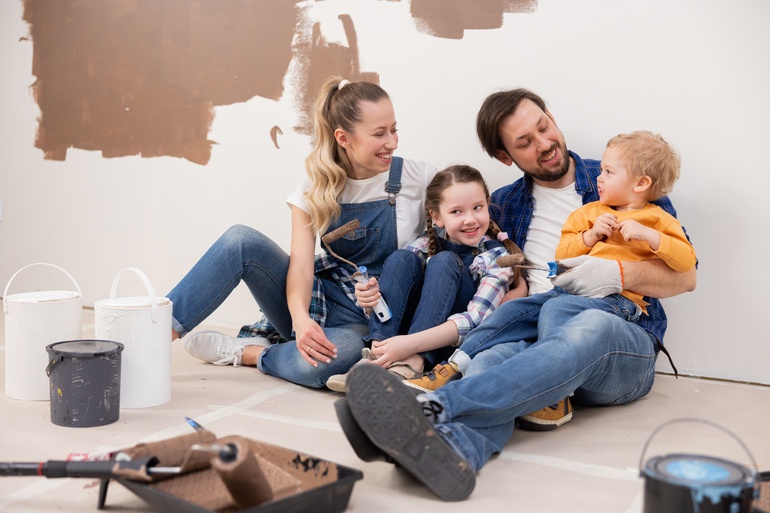 Two parents with their kids in their home with cans of paint and a roller in front of them during a wall painting project.