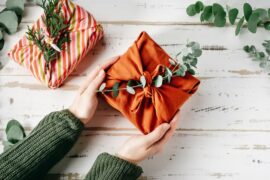 An overhead view of hands holding an orange, fabric-wrapped gift. Another fabric-wrapped gift sits on the table.