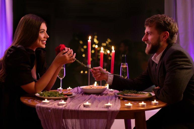 A couple is sitting across from one another, smiling, at a candlelit dinner. The man is giving the woman a rose.