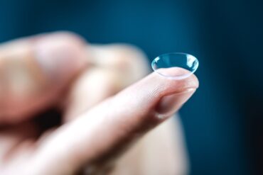A close-up of a finger holding a clear contact lens. The background and the person's hand are blurred.