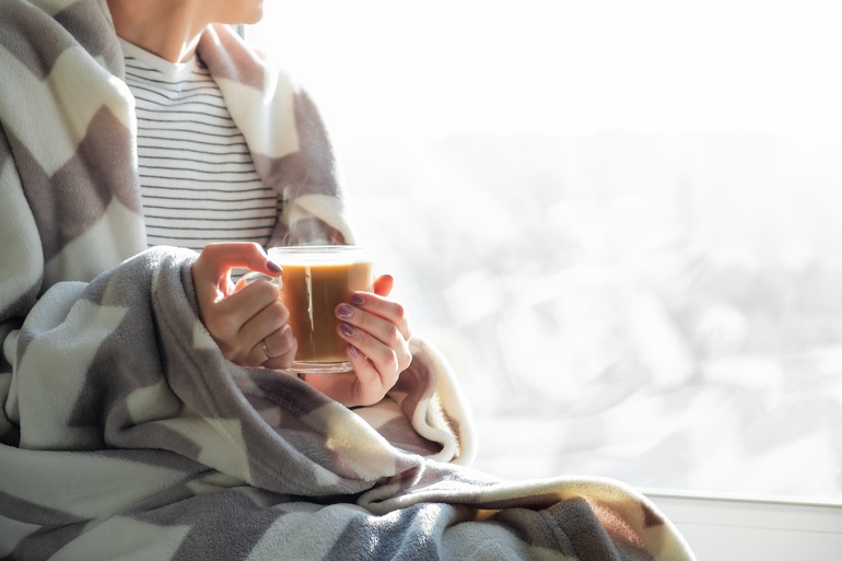 A close-up view shows a person holding a clear mug filled with coffee. The person has a blanket wrapped around them.