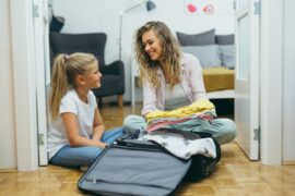A smiling woman and young girl sort through clothes to pack into a suitcase. Both are seated on wooden flooring.
