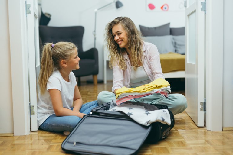 A smiling woman and young girl sort through clothes to pack into a suitcase. Both are seated on wooden flooring.