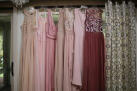 A selection of hanging bridesmaid dresses in different colors, styles, and fabrics. There's a curtain to the right.