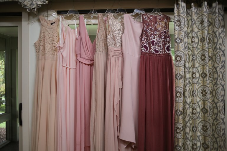 A selection of hanging bridesmaid dresses in different colors, styles, and fabrics. There's a curtain to the right.