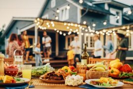 An outdoor table covered in food and drink. People gather in the background, talking beneath glowing lights.