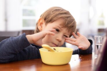 A young boy at the dinner table rubbing his eyes as he appears frustrated by his meal. There's a yellow bowl in front of him.