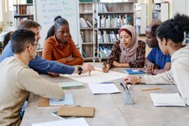 A group of diverse college students sitting at a library table to discuss a project. They're working off one large document.