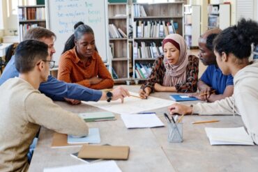 A group of diverse college students sitting at a library table to discuss a project. They're working off one large document.