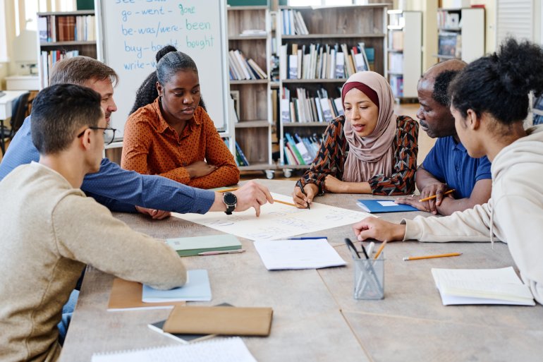 A group of diverse college students sitting at a library table to discuss a project. They're working off one large document.