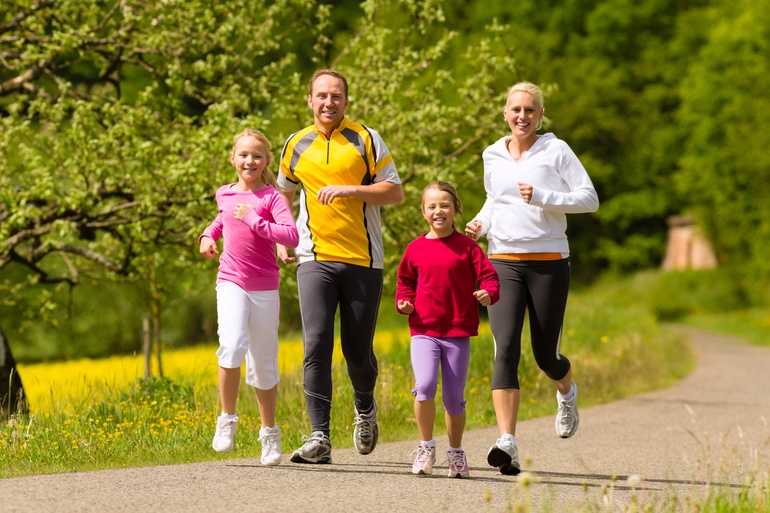 A family of four running together along a trail surrounded by a beautiful meadow. It's sunny outside and they're smiling.