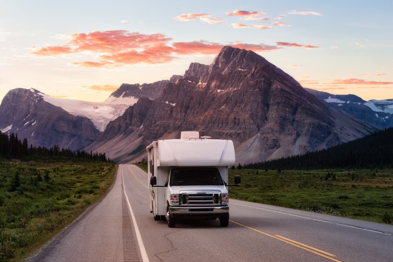 An RV on the road driving out of the mountains and away from the sunset behind it. There are pink and orange clouds.