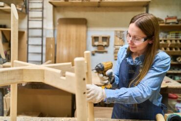 A woman in overalls, a work shirt, and gloves uses a power drill on a wooden structure in a woodworking shop.