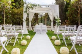 An outdoor wedding venue with all-white decor, a white aisle and chairs, and a large canopy with flowers.
