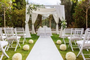An outdoor wedding venue with all-white decor, a white aisle and chairs, and a large canopy with flowers.