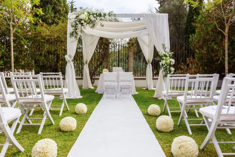 An outdoor wedding venue with all-white decor, a white aisle and chairs, and a large canopy with flowers.