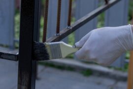 A close-up view shows a person's hand wearing a white latex glove as they use a paintbrush to paint a metal railing.