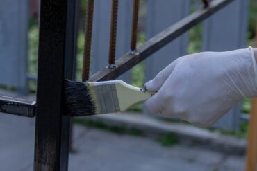 A close-up view shows a person's hand wearing a white latex glove as they use a paintbrush to paint a metal railing.