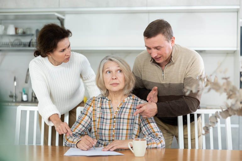 An older woman sitting at a table, signing a paper, with a man and a woman arguing over her shoulders.