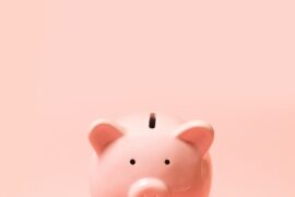A simple pink ceramic piggy bank with two black dots for eyes. It is positioned against a similarly-colored background.