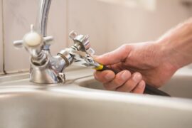 An close-up view of a person's hand using black pliers to twist and fix a silver-plated tap in the sink.