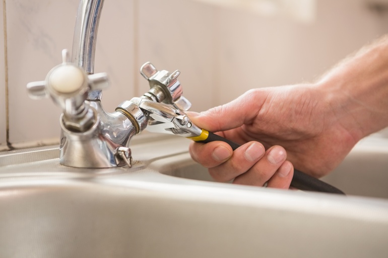 An close-up view of a person's hand using black pliers to twist and fix a silver-plated tap in the sink.