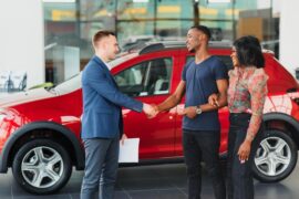 In front of a red car, a man wearing a blue jacket shakes the hand of another man who is standing next to a woman.