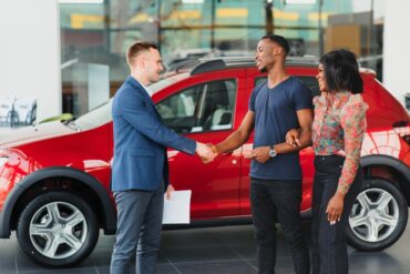 In front of a red car, a man wearing a blue jacket shakes the hand of another man who is standing next to a woman.