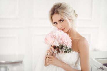 A blonde woman wearing a strapless wedding dress holds a pink bouquet close to her face and stands in an all-white room.