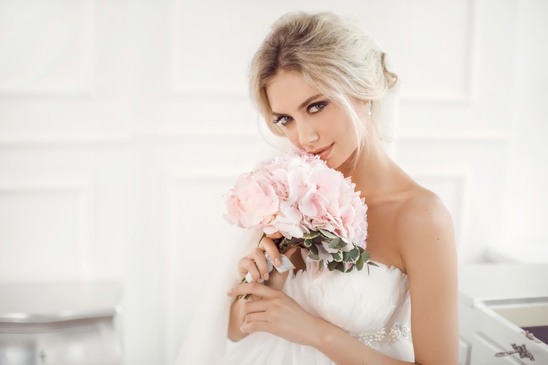 A blonde woman wearing a strapless wedding dress holds a pink bouquet close to her face and stands in an all-white room.