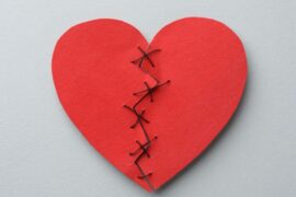 A red heart, once ripped in two pieces, sewed back in one piece with thread, against a plain light gray background.