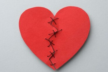 A red heart, once ripped in two pieces, sewed back in one piece with thread, against a plain light gray background.