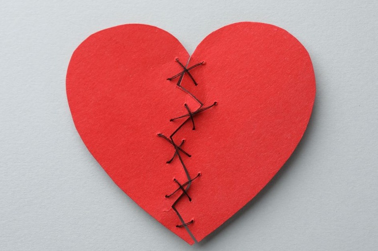 A red heart, once ripped in two pieces, sewed back in one piece with thread, against a plain light gray background.