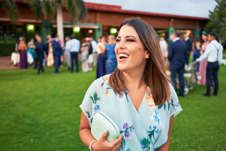 A close-up of a smiling woman wearing a floral dress and carrying a purse. Wedding guests stand behind her, mingling.