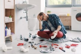 A man sits on a bathroom floor with several tools surrounding him. He holds his head with a frustrated expression.