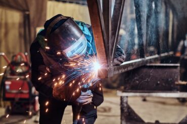 A fabrication professional wearing a helmet and uniform as they weld together pieces of metal. Sparks are flying.