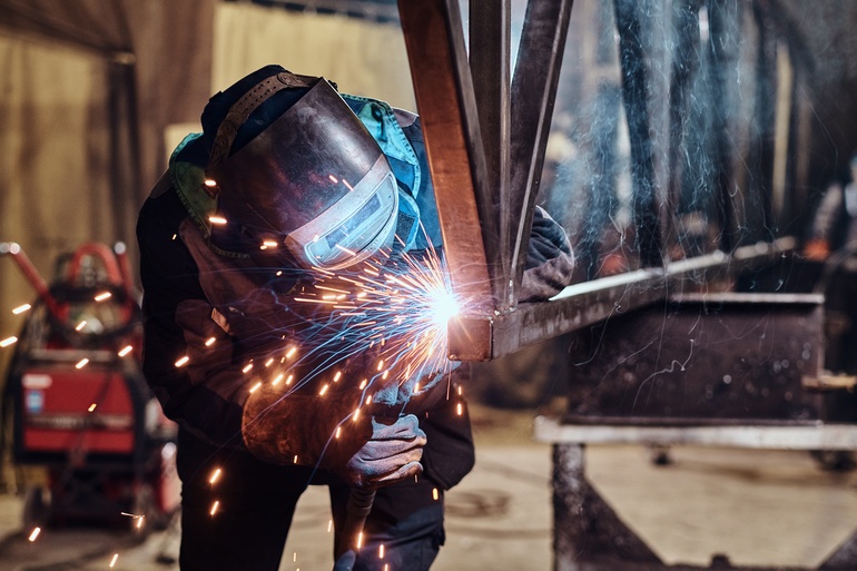 A fabrication professional wearing a helmet and uniform as they weld together pieces of metal. Sparks are flying.