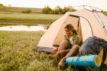 A woman ties her boots while sitting inside a beige tent near a body of water with a backpack and teal mat within reach.