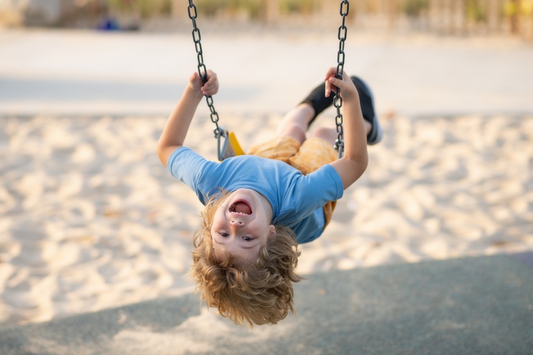 A joyous child hanging his head back as he swings at a park. Sand covers the ground beneath him.