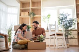 Woman and man sitting on wooden floor inside an apartment, looking at a piece of paper, with multiple plants around.