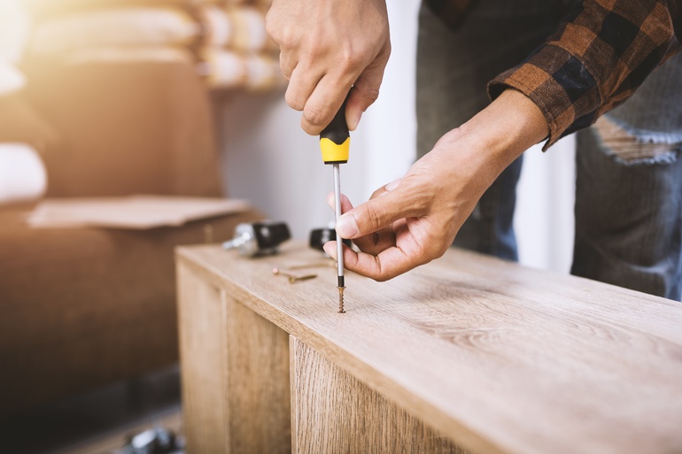 A close-up of a person in a plaid shirt using a yellow screwdriver to assemble a piece of wooden furniture.