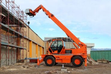 An orange telehandler sits with its arm up near a commercial building that's under construction and covered with scaffolding.