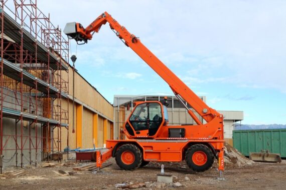 An orange telehandler sits with its arm up near a commercial building that's under construction and covered with scaffolding.