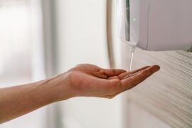 Close-up of a person placing their hand under a hand sanitizer gel dispenser. It dispenses some into their palm.