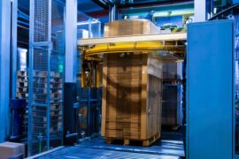 A large, blue stretch wrap machine is wrapping a pallet with flat cardboard boxes with stretch film.