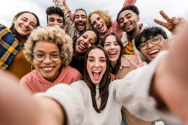 A group of friends all crowd around each other and take a selfie together, smiling with the clouds behind them.